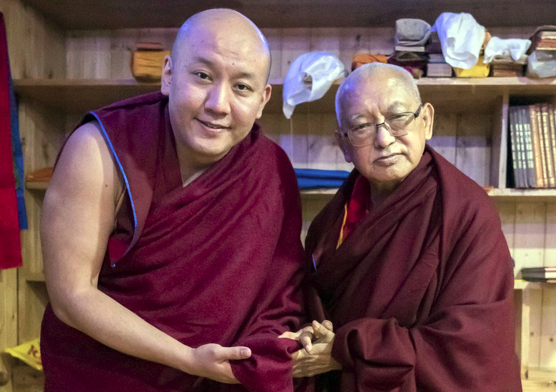 Zong Rinpoche and Lama Zopa Rinpoche holding hands and posing for the camera in front of bookshelves containing Dharma items inside at Tushita Meditation Center in Dharamsala