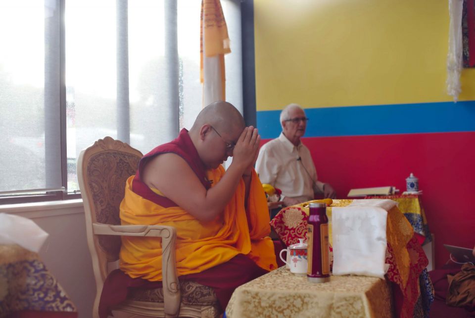 His Eminence Ling Rinpoche with hands folded together and held at Rinpoche's forehead seated on a chair at the front of a colorful room at shantideva center.