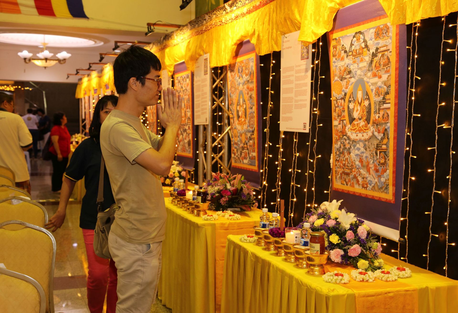 People standing in front of thankgas of Lama Tsongkhapa hung on a wall with tables covered in offerings below them.