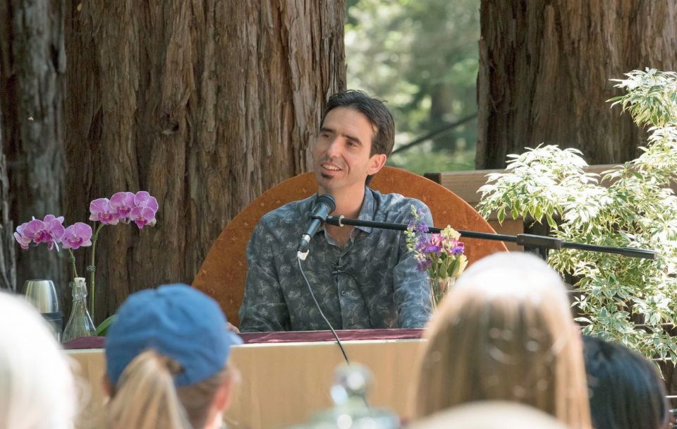 Tenzin Ösel Hita seated outdoors in front of beautiful old trees in the woods smiling at an audience listening to him talk.