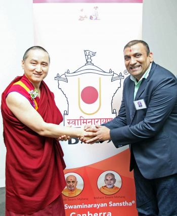 Geshe Tenzin Zopa and Mr. Dipak Gondaliya have grasped each others hands and are smiling upon meeting each other while standing in front of a sign with the name BAPS Swaminarayan Sanstha on it.