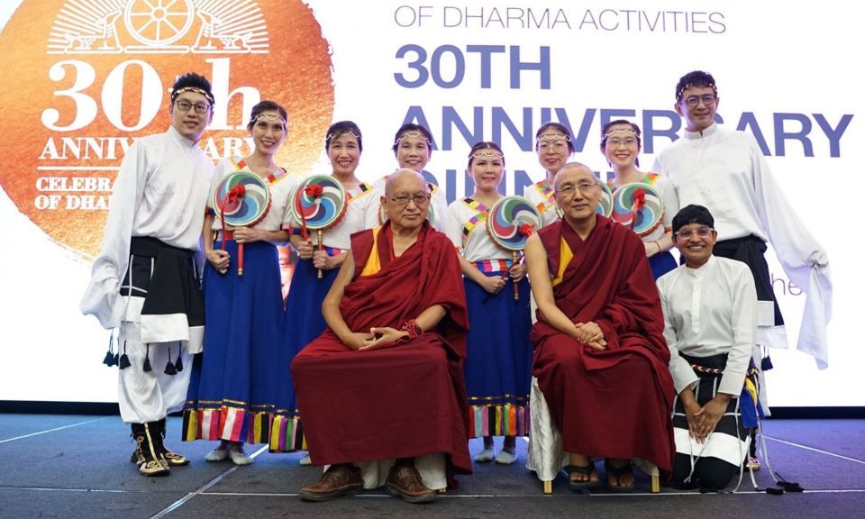 A group of dancers wearing mostly white traditional Tibetan styled dress, with the women in bright blue long skirts posing for a photo with Lama Zopa Rinpoche and Khen Rinpoche Geshe Chonyi on stage with the large 30th anniversary banner hanging up behind them.