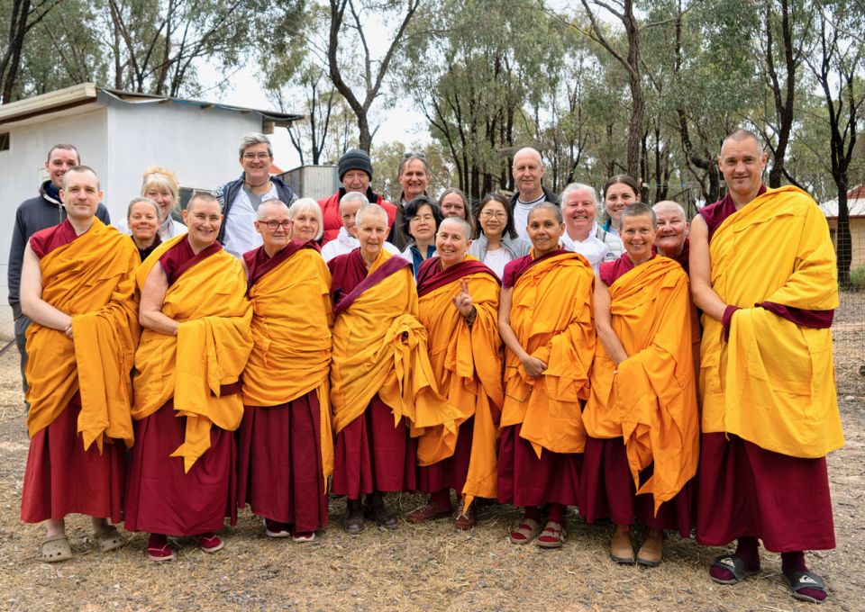 Group of people standing together with half of the group western monastics standing outside together smiling and posing for the camera with the nun in the middle of the front row making the peace sign with one hand.