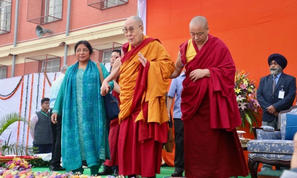 Doctor Renuka Singh standing next to and holding the hand of His Holiness the Dalai Lama while an attendant holds His Holiness's other arm on stage at the celebration.
