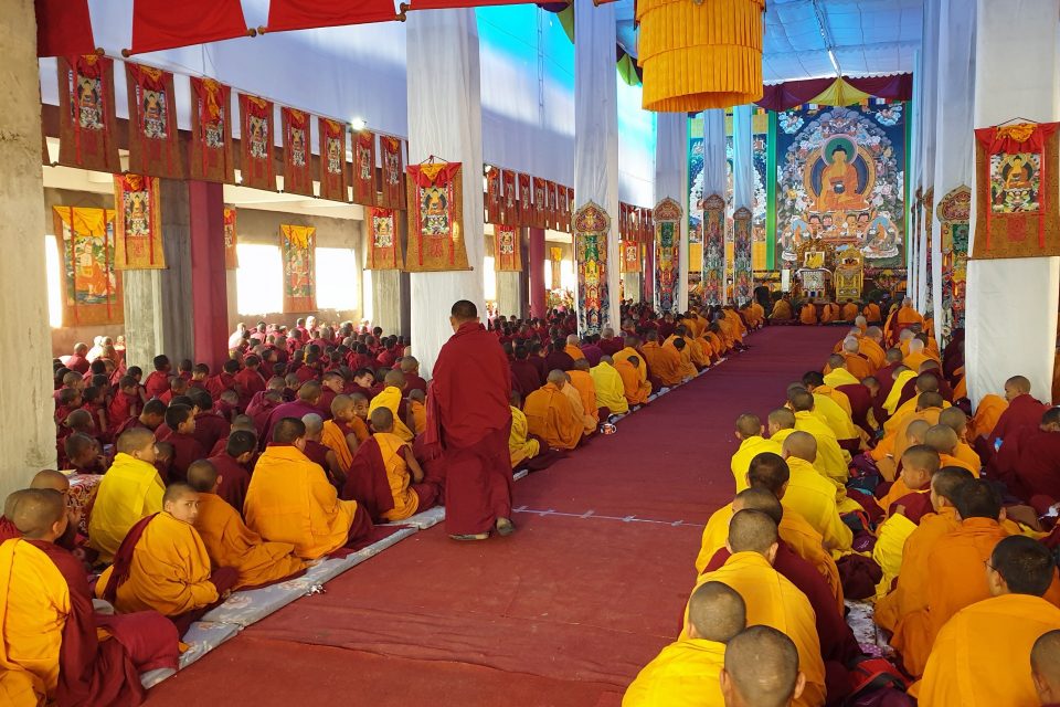 A room full of monks and nuns seated in rows facing the front of the room where there is a large image of the Buddha.