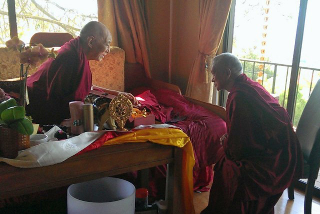 A smiling Choden Rinpoche seated on his bed with a smiling Lama Zopa seated in a chair and leaning towards Choden Rinpoche.