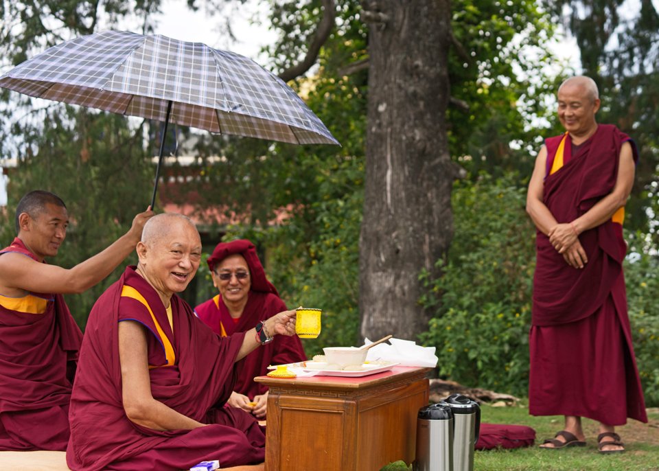 Lama Zopa Rinpoche holding up a tea mug sitting under tree