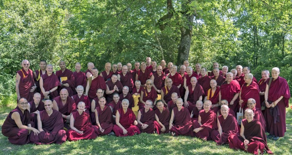 Lama Zopa Rinpoche and a large group of monks and nuns smiling together underneath tall trees.