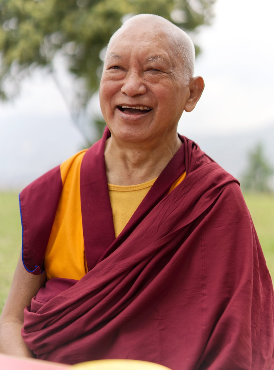 Lama Zopa Rinpoche smiling outside with a tree in the background