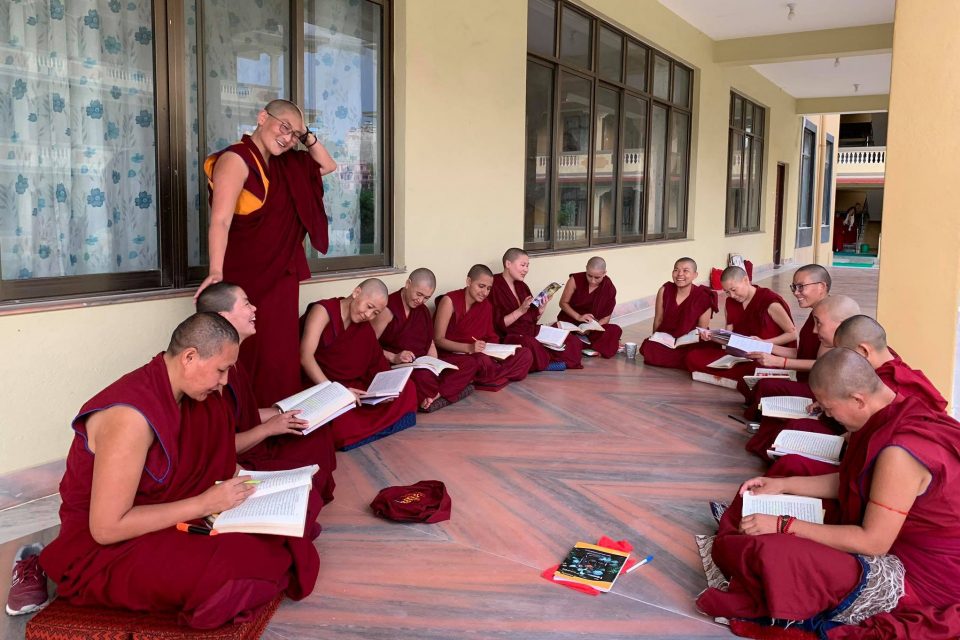 A group of nuns seated in a circle smiling with books open in their laps and Geshema Namdrol Phuntsok standing and smiling.