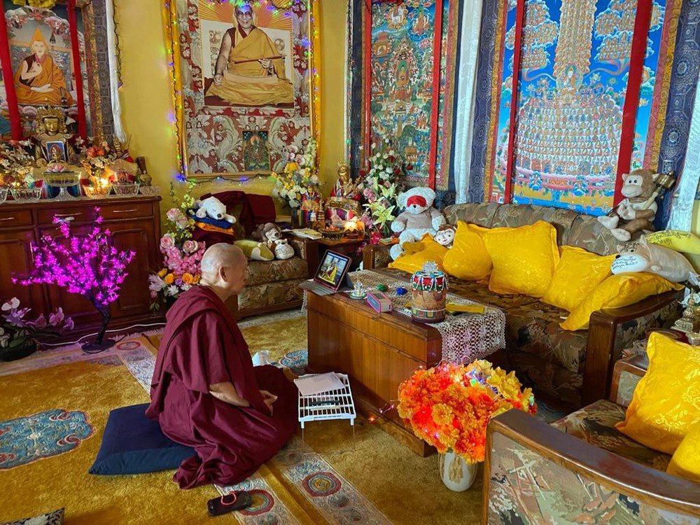 Lama Zopa Rinpoche seated in front of a laptop watching His Holiness with a big thankga of His Holiness hanging on the wall in the room background.