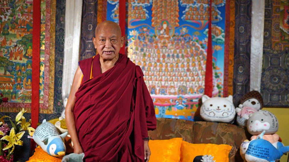 Lama Zopa Rinpoche standing in front of a thangka of the merit field and a couch from which he gives his video teachings