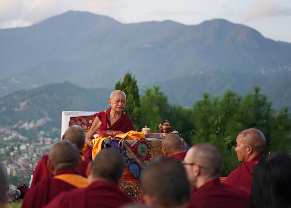 Lama Zopa Rinpoche sitting on a low throne outside with hills and mountains in background