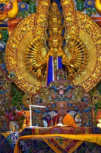 Lama Zopa Rinpoche on teaching throne with hands in prostration mudra with 1000-Arm Chenrezig statue behind him