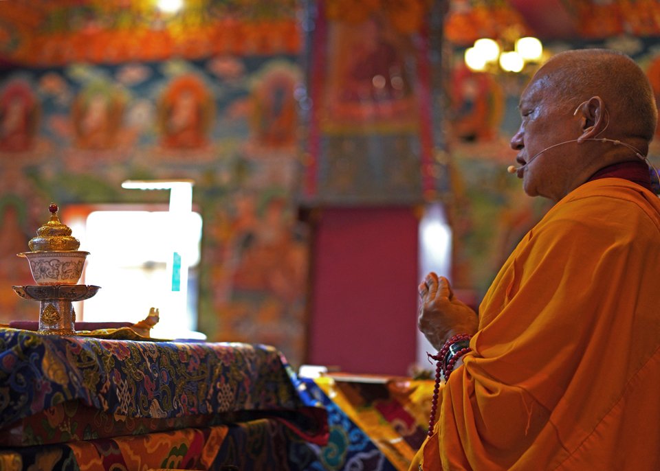 Profile view of Lama Zopa Rinpoche with hands together in prostration mudra on teaching throne