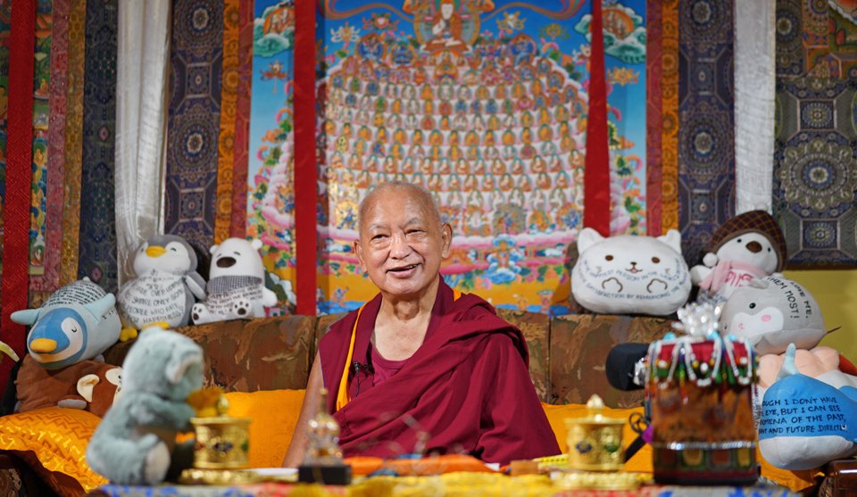 Lama Zopa Rinpoche seated on a couch in front of a thangka of the merit field and surround by stuffed toyes with mantras and Dharma messages written on them