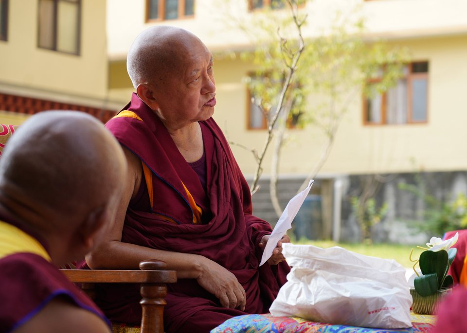 Lama Zopa Rinpoche sitting outside, talking and holding a piece of paper with nunnery buildings in background