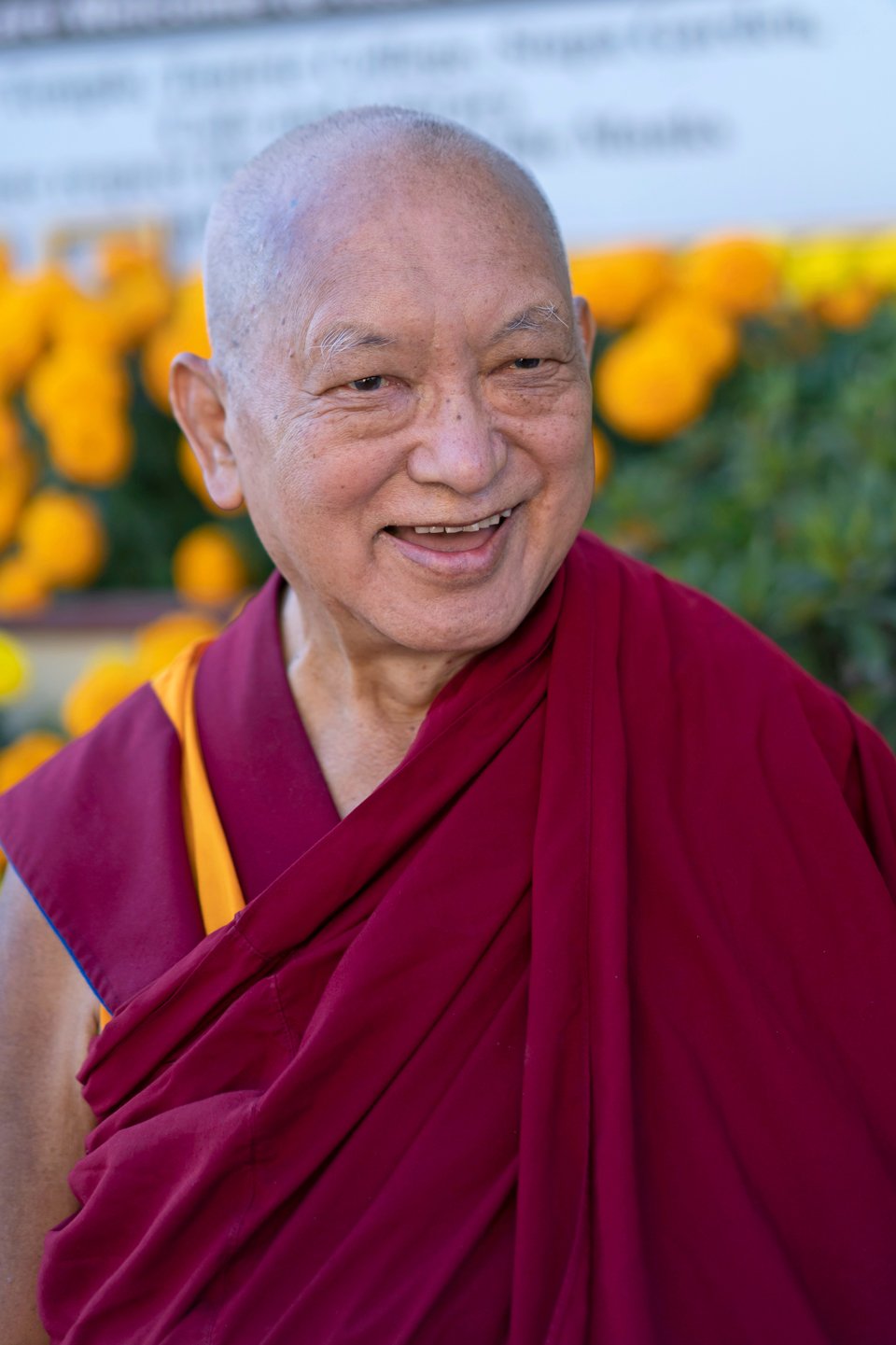 Lama Zopa Rinpoche smiling outside with blooming marigolds behind hime