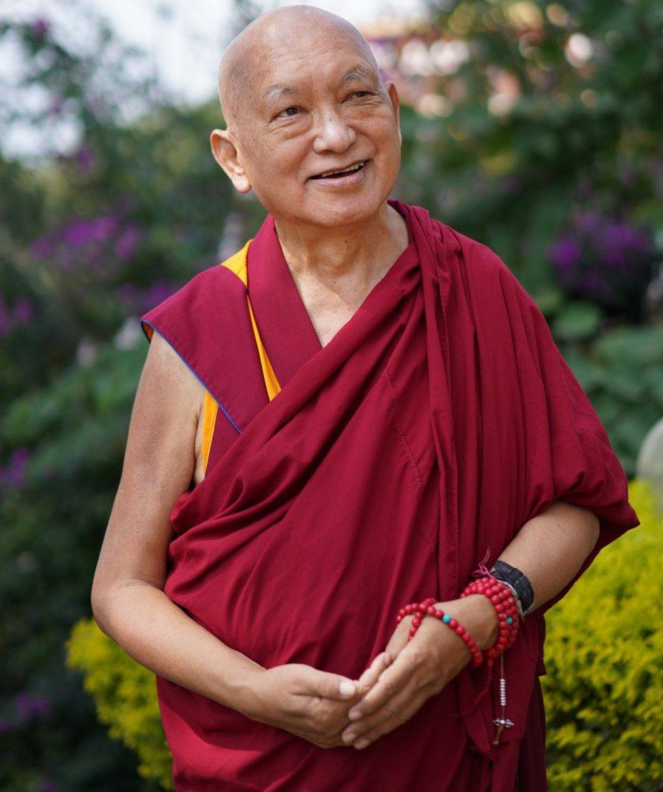 Lama Zopa Rinpoche smiling in a garden with green shrubs and purple blossoms behind him in the background