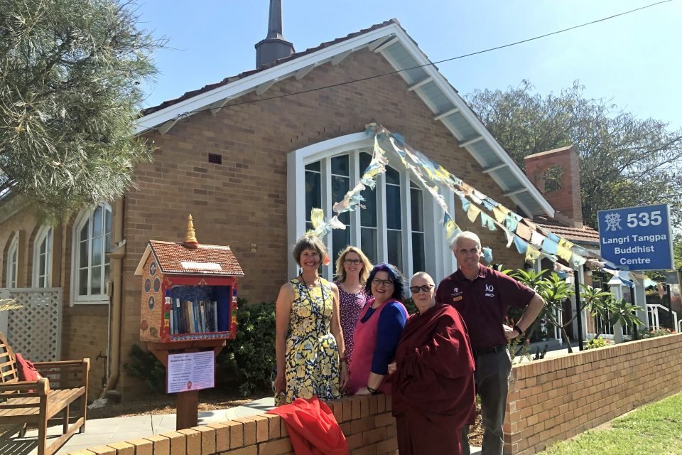 A group of smiling people standing in front of the new street library with the converted church dharma center behind them.