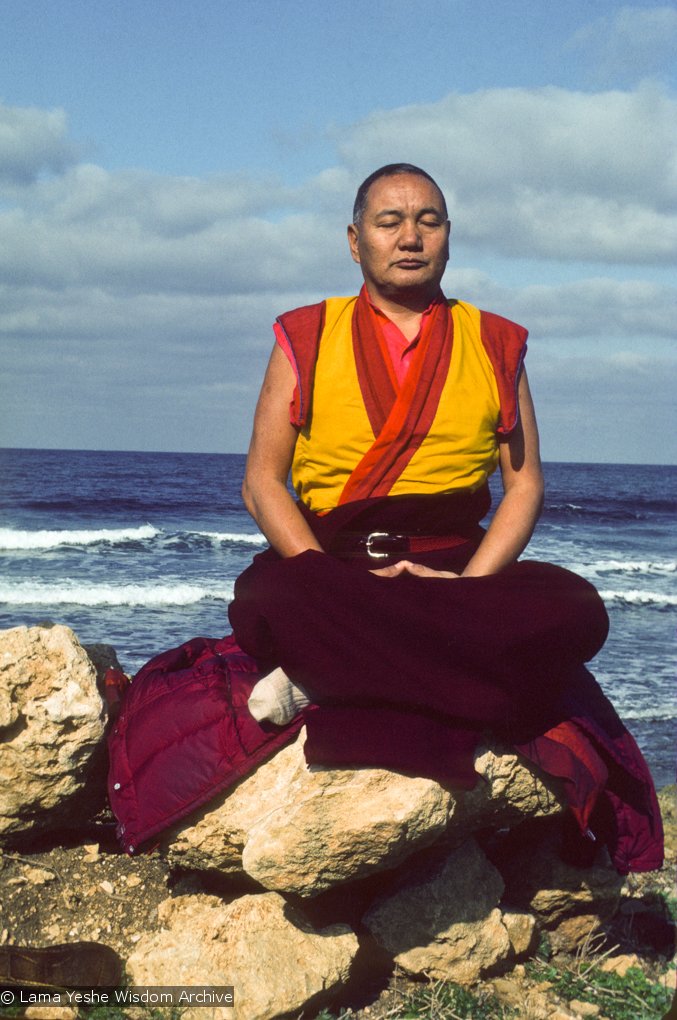 Lama Yeshe, with eyes closed and seated on a rock, meditating next to the sea
