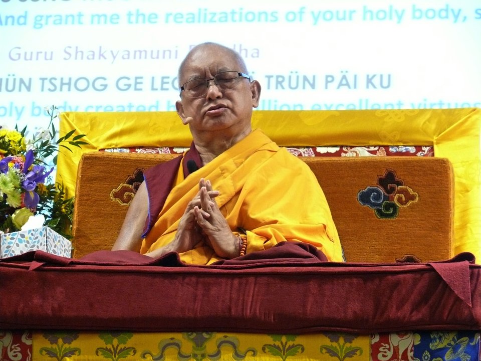 Lama Zopa Rinpoche on the teaching throne with prayers projected on screen behind him