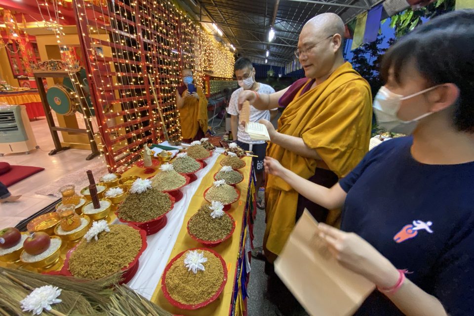 Two monastics and two lay people wearing face masks standing in front of a row of bowls of different grains and making offerings.