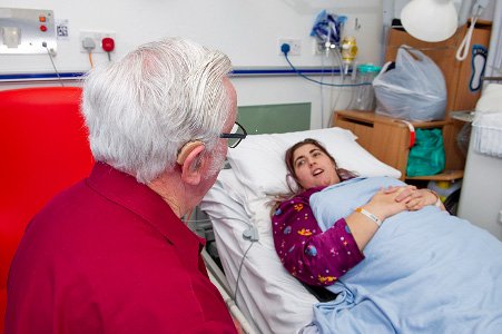 Chaplain seated beside a young person lying on their back on a hospital style bed in a hospital style room.