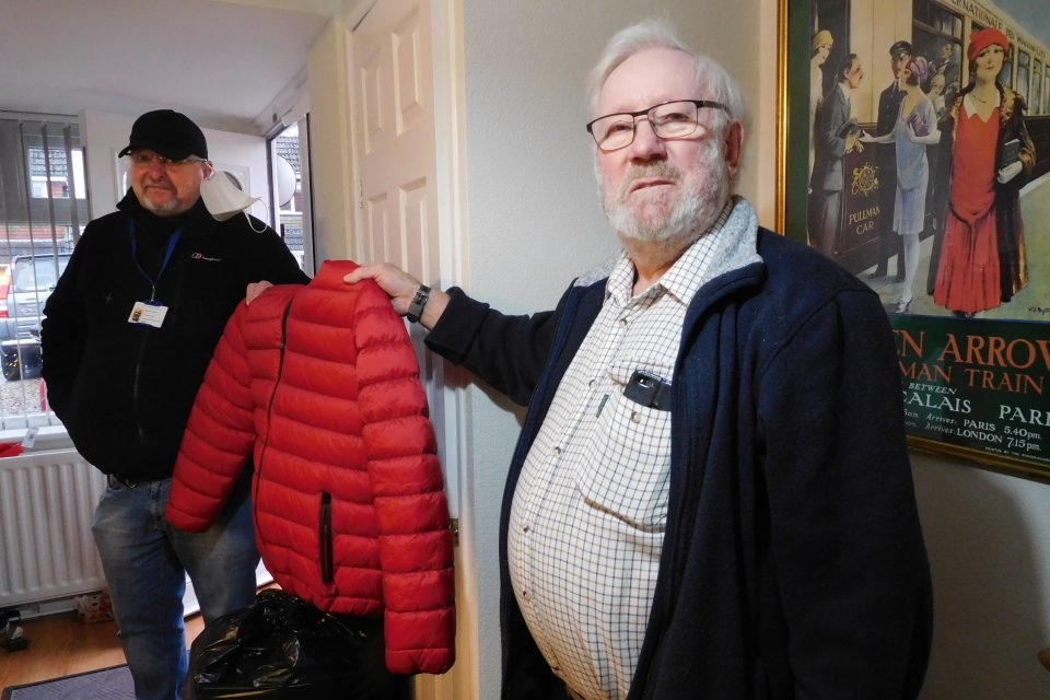 Chaplain and person wearing a name badge and face mask together holding up holding up a red winter jacket.