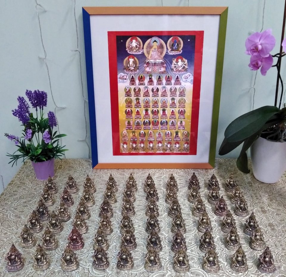 Six rows of small bronze colored statues neatly arranged on a tablecloth in front of a framed picture of Buddhas.