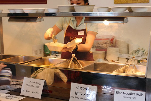 Person wearing an apron putting a serving of food into a container while standing behind trays of coconut milk rice and other foods that have signs with the names of the foods and prices in front of them.
