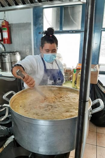A person wearing a face mask standing in a kitchen over a gigantic pot stirring beans.