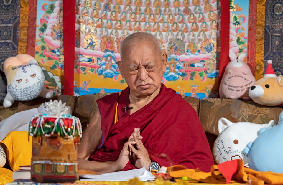 rinpoche with eyes closed and hands in prostration mudra sitting on couch with stuffed toys and a large merit field thangka behind him