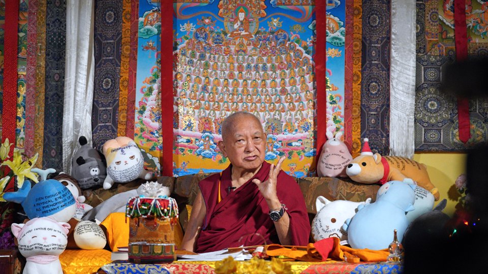 Lama Zopa Rinpoche seated on a couch, surrounded by stuffed toys with Dhamra messages written on them, gesturing with his hand while teaching