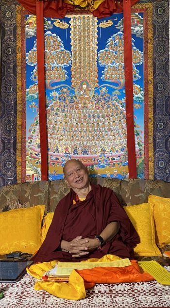 Rinpoche smiling on his couch seating below a large thangka of the merit field