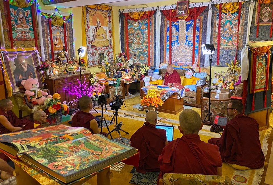 Lama Zopa Rinpoche sitting under lights in his room teaching while students listen and the video camera records