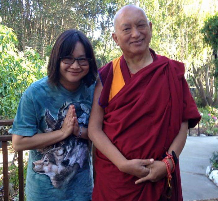 Person with hands folded in prostration standing next to Lama Zopa Rinpoche with both smiling outdoors in front of trees.