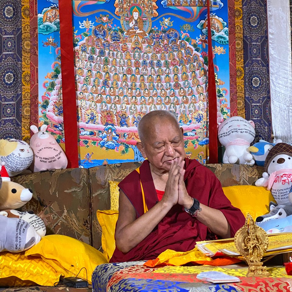 Lama Zopa Rinpoche with hands together in prostration mudra seated in front of large merit field thangka
