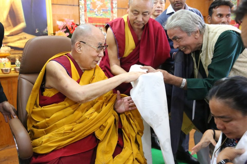 The Dalai Lama accepting a white scarf from a smiling person.