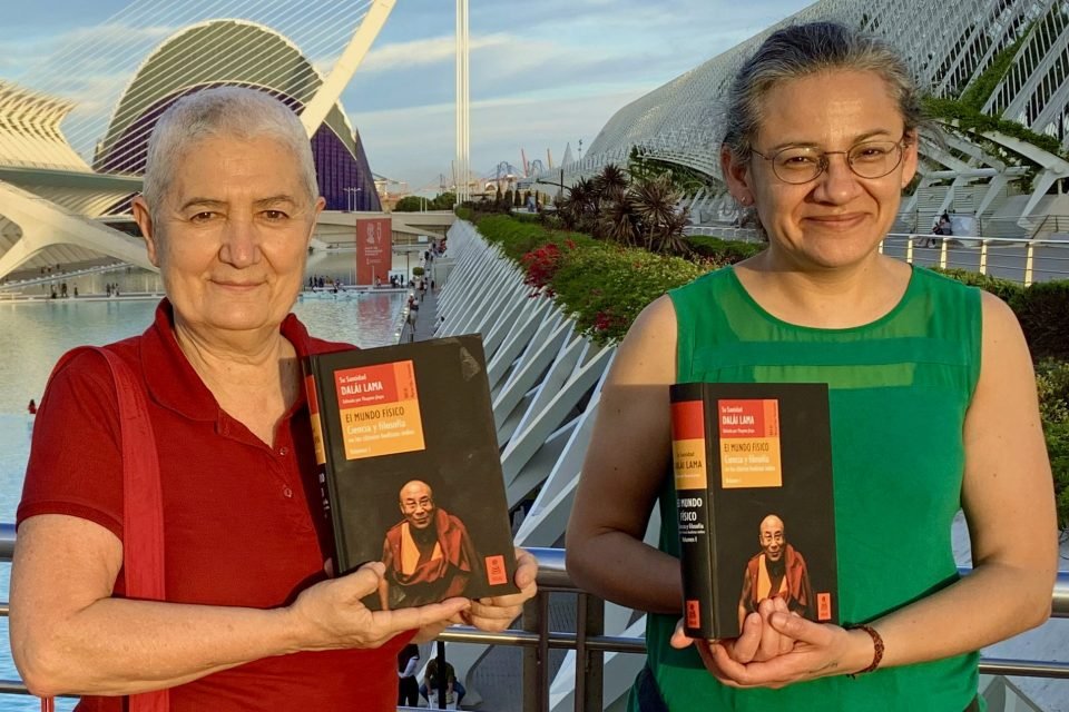 Two people holding up one book each with a picture of the Dalai Lama on the cover while standing in front of a beautiful landscape.