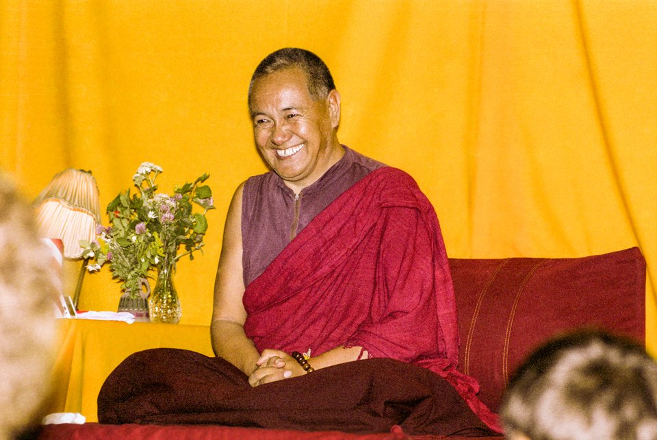 Lama Yeshe smiling at the audience, while seated on low throne in front of gold curtain