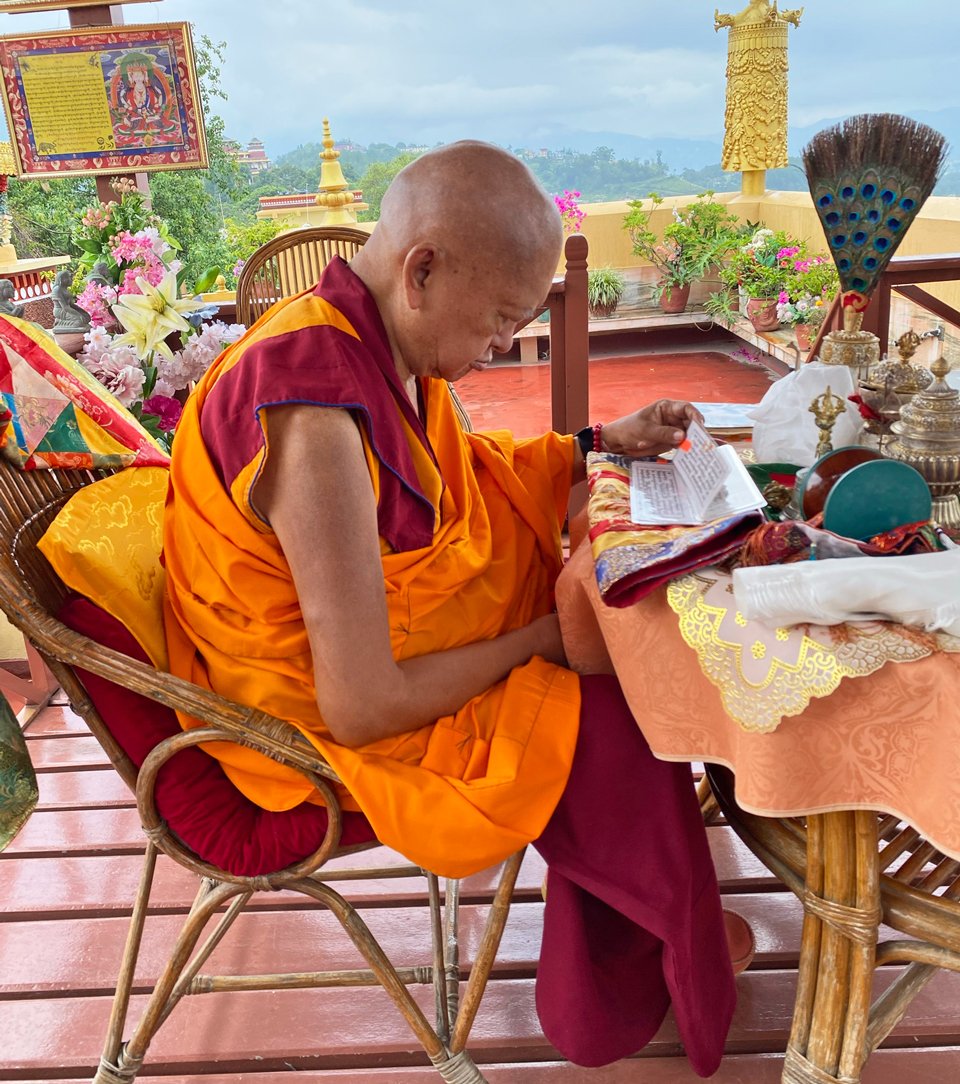 Lama Zopa Rinpoche seated at a covered roof-top patio table with texts and ritual implements and foothills in the background