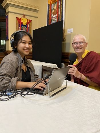 Two people seated at a table with a laptop microphone and headset recording a podcast.