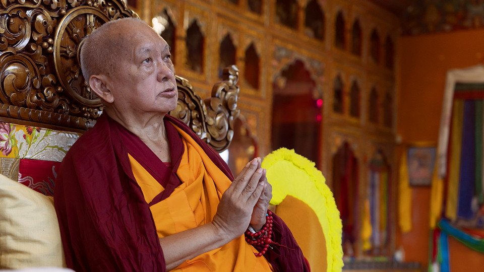 Lama Zopa Rinpoche with hands together on a throne in a gompa with a yellow puja hat next to him