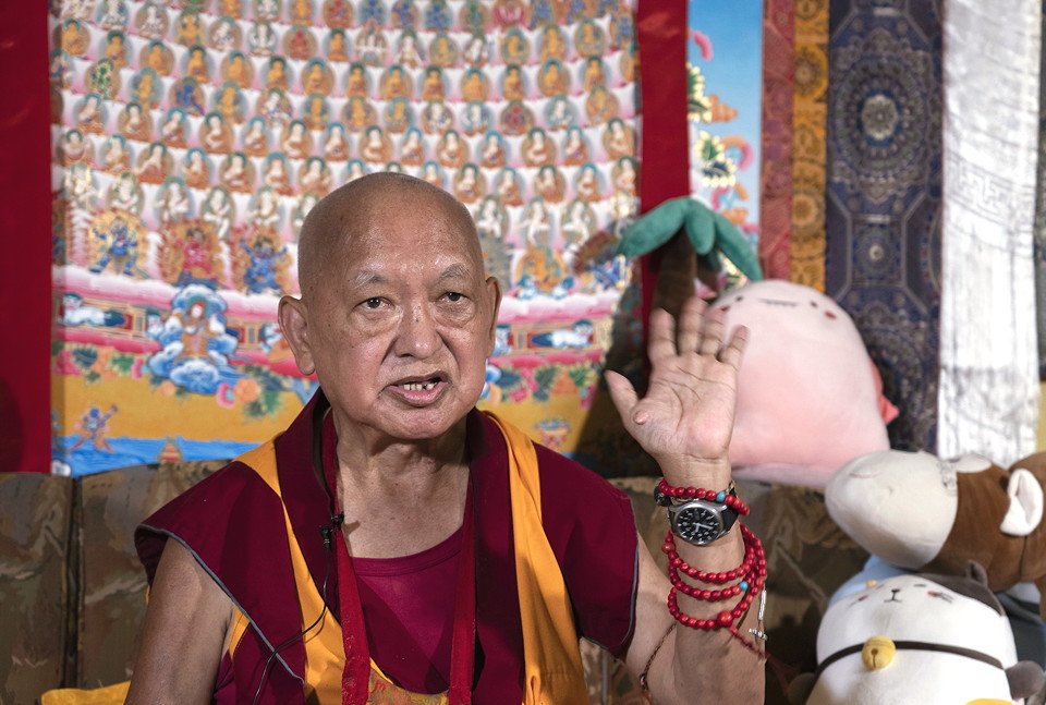 Lama Zopa Rinpoche seated on a couch with a Merit Field thangka and stuff animals behind him