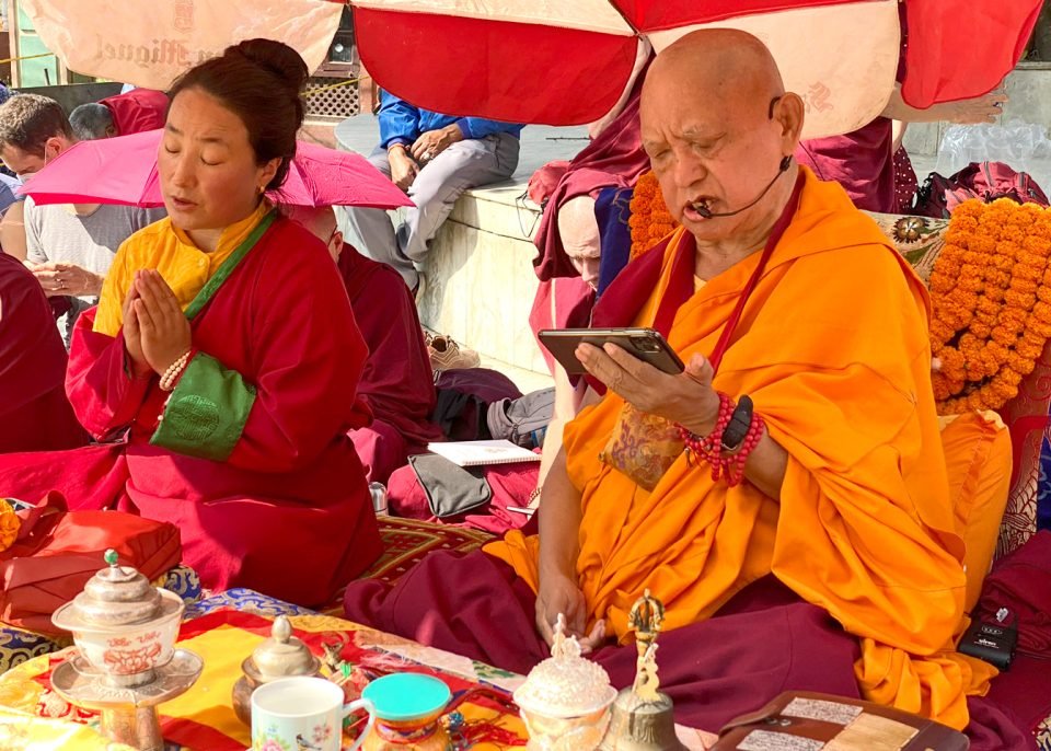 Lama Zopa Rinpoche and Khadro-la sitting under an umbrella doing prayers with a puja table in front of them 