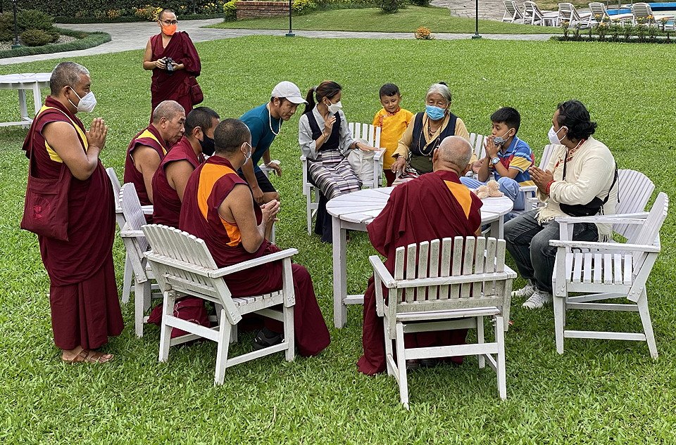 a group of monks and family members sitting around a white table with Lama Zopa Rinpoche on a green lawn