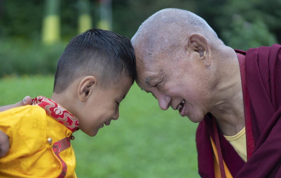 Lama Zopa Rinpoche touching foreheads with a smiling young boy with a green lawn in the background