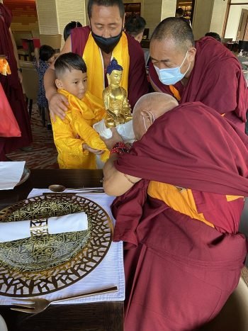 Lama Zopa Rinpoche bowing and offering a golden statue of Buddha to a young boy in gold shirt