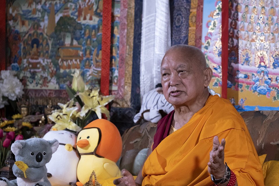 Lama Zopa Rinpoche seated on a couch with stuffed animals and thangkhas behind him